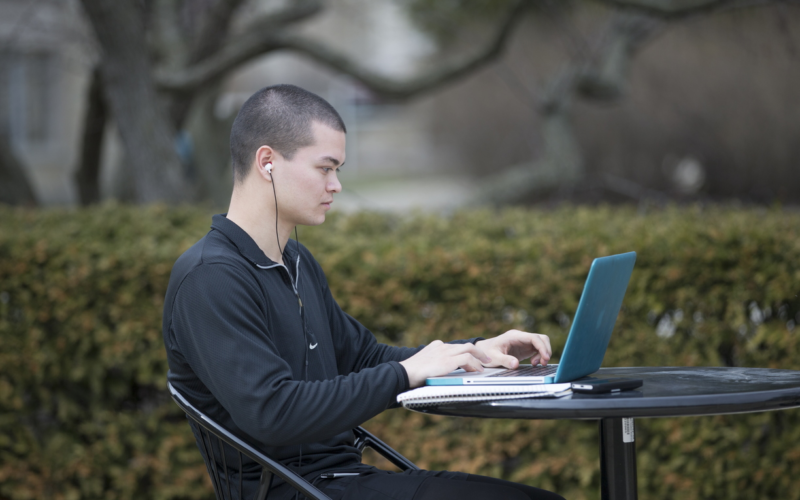 Male student sitting at picnic table outside typing on laptop computer with earbuds in ears