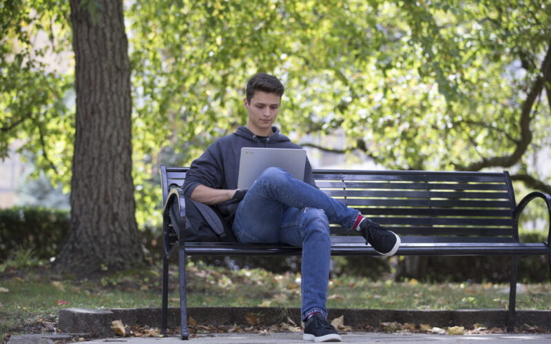 student sitting on park bench looking at laptop