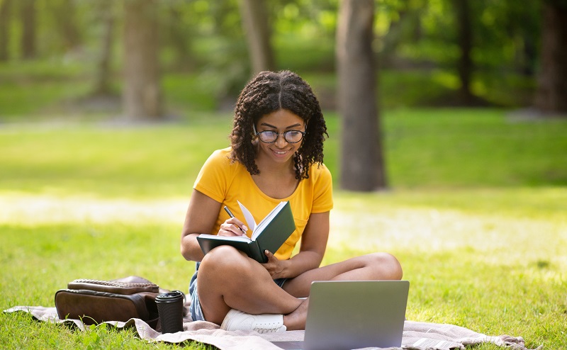 Black woman writing in notebook near laptop computer at park