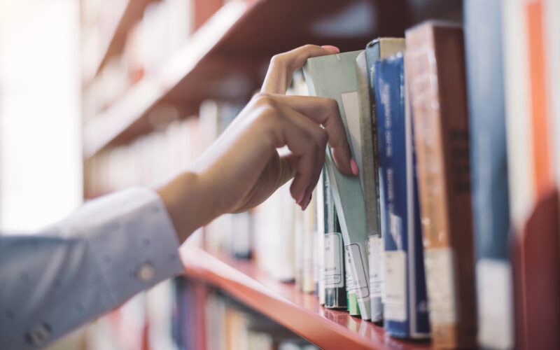 Woman at the library, she is searching books on the bookshelf and picking a textbook, hand close up