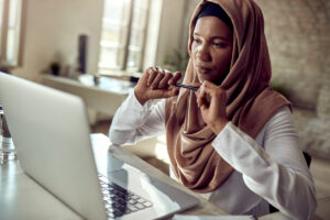 Black Muslim female professor looking at laptop screen pensively and holding pen in hands