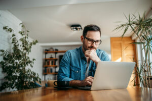 White man wearing glasses looking at laptop screen and thinking with hand on chin