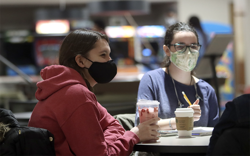 two students wearing masks working at a table and drinking beverages