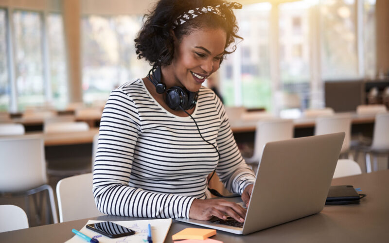 Smiling college student sitting on campus wearing headphones and working online with a laptop