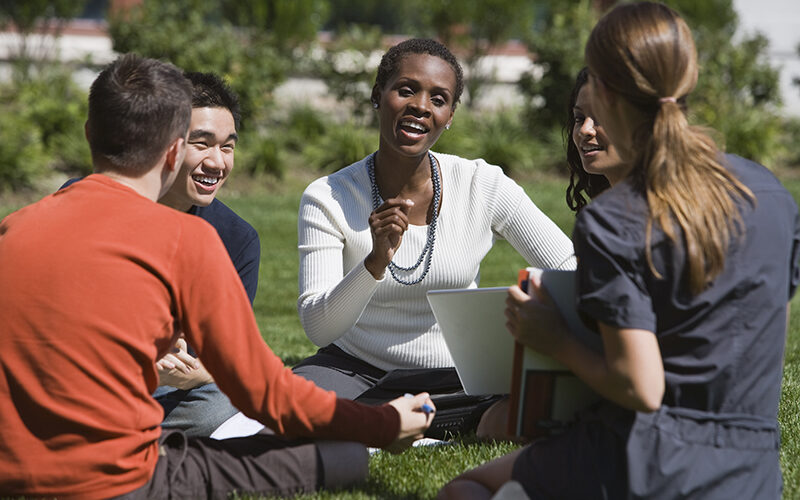 four students with professor sitting in a circle outside on grass, discussing