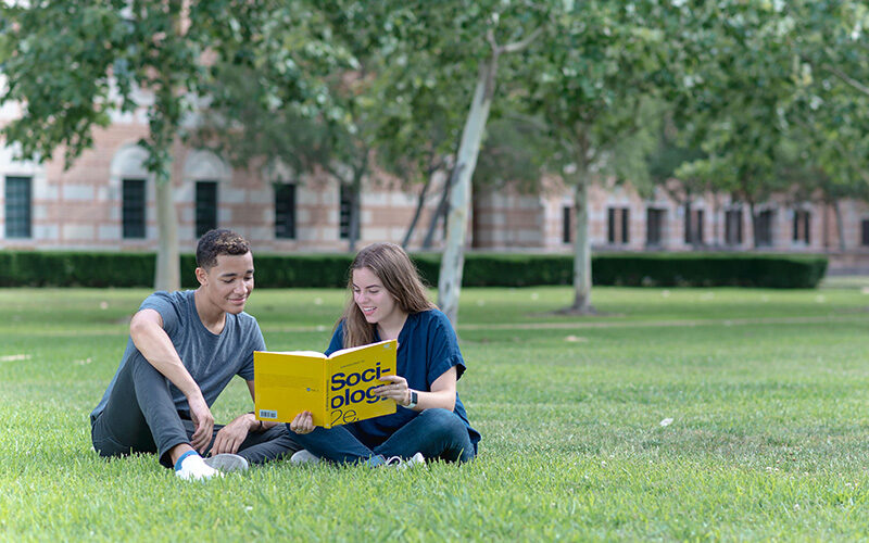 two students sitting on grass reading textbook