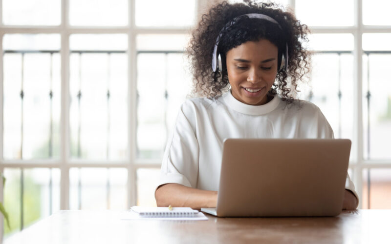 African American woman smiling while typing on laptop and wearing headphones.