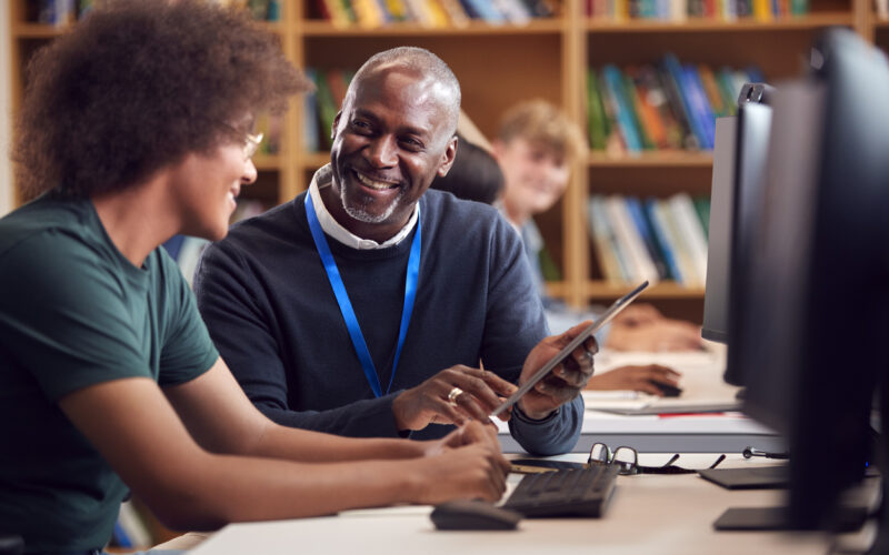 Smiling black professor helping black male student sitting at computer in library