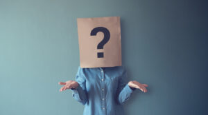 Person with arms up questioning pose wearing brown bag over head with question mark on it.