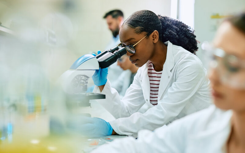 Black woman scientist looking into microscope with another woman scientist in the foreground and a male scientist in the background.