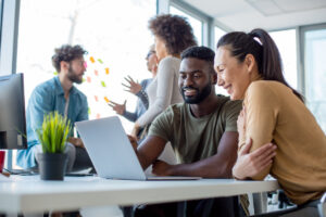 Students sitting at a table looking at a laptop and smiling with a group of students talking to each other in the background. 