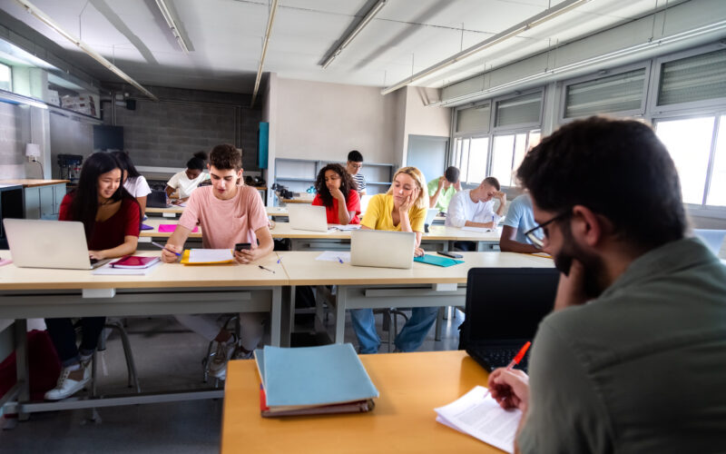 Male teacher marking exams with red pen while students look bored at their tables in the classroom.