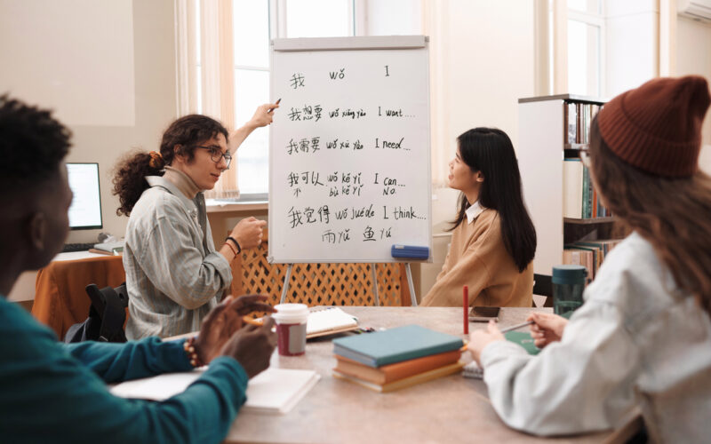 Group of students around a table looking at a whiteboard with beginning Chinese language writing on it.