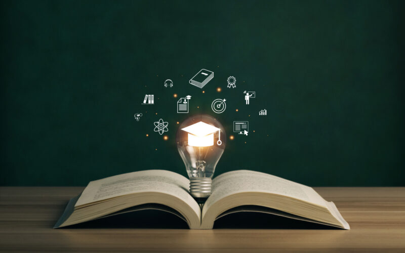 Book open on table with lightbulb lit up with mortarboard cap illuminated inside and icons representing "knowledge" floating around it
