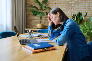 Tired woman sitting at desk with laptop and textbooks, resting her head on her hand