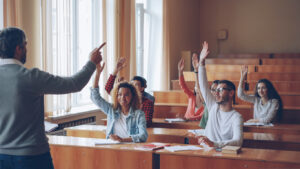 Group of students sitting in classroom, listening to professor, smiling and raising hands. 