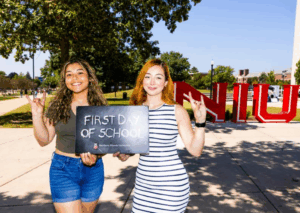 Two female presenting students holding first day of school sign in front of NIU letters