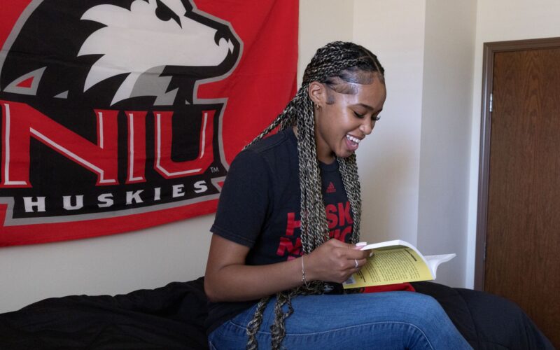 Female student sitting on dorm bed reading a book with an NIU Huskies flag on the wall behind her