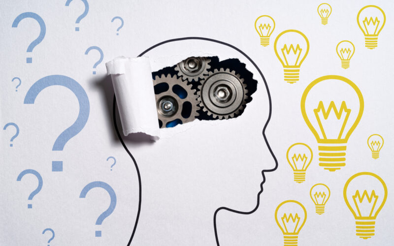 Outline drawing of head with question marks and lightbulbs drawn around it. Paper torn at top of head to reveal metal gears as "brain."