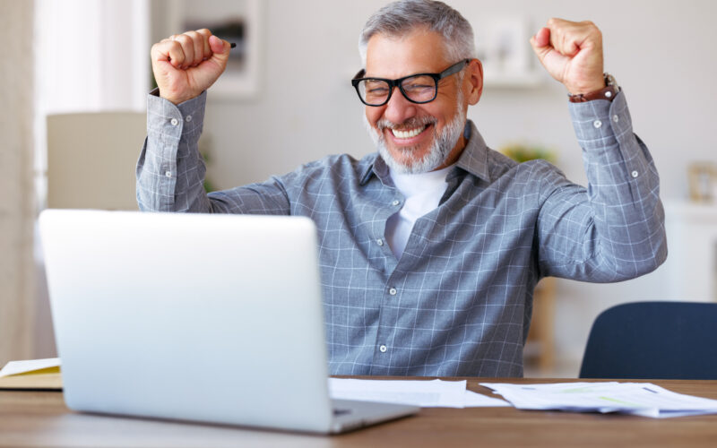 Excited older man raising fists in celebration and looking at laptop screen.