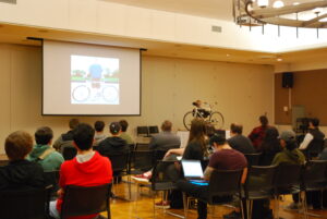 Dr. Brian Coller holding up a bicycle in front of room of seminar attendees