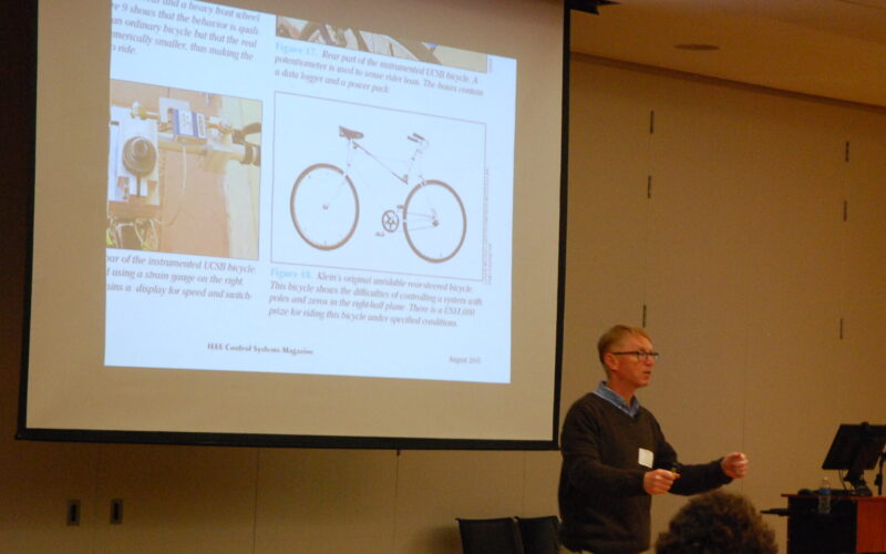 Dr. Brian Coller standing in front of projector screen with bicycle image on it