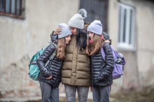 Woman kisses her daughter on forehead, has arms around both daughters, outside, trauma