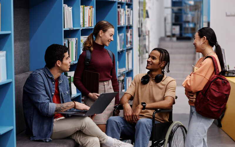 Diverse group of students including young man in wheelchair chatting in college library