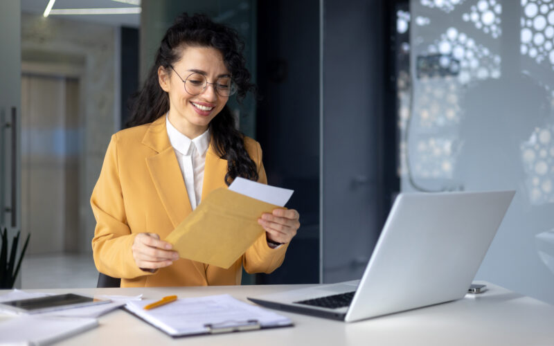 Hispanic woman smiling at envelope in office with laptop and papers on desk in front of her