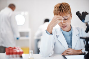 Female scientist in a white lab coat sits at a laboratory desk, resting a hand against the forehead in a gesture of fatigue or stress
