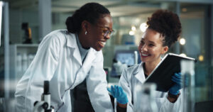 Two smiling female scientists in lab coats collaborate in a laboratory, with one holding a tablet and wearing blue gloves while the other leans forward near a microscope.