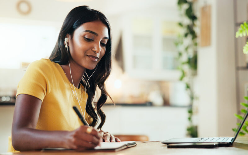 A woman wearing a yellow short-sleeve top is seated at a wooden table, writing in a notebook with a pen. Earphones are connected to a laptop placed on the table.