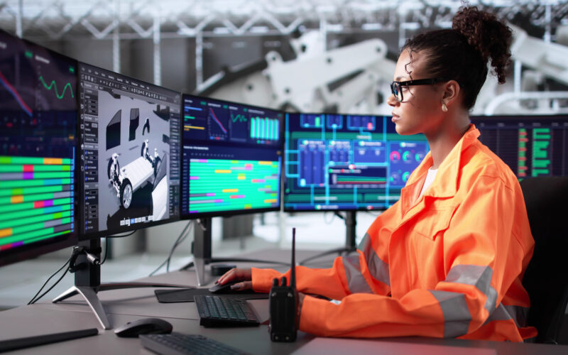 A female engineer wearing a bright orange safety jacket sits at a workstation with multiple large monitors displaying technical data, charts, and a 3D model of a vehicle chassis. The engineer is using a keyboard and mouse, with a two-way radio placed on the desk. The background shows an industrial setting with machinery and equipment.