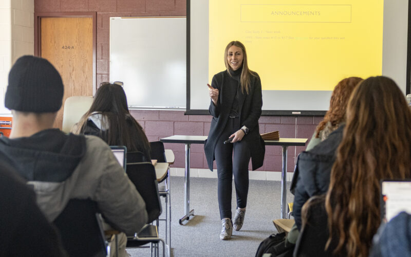 Prof. Stephanie Baker leans on front of table at the front of classroom with students sitting at desks.
