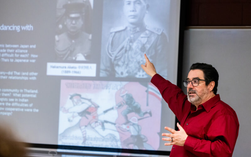 E. Taylor Atkins, in a red shirt, stands at the front of a classroom, gesturing and pointing toward a large projected slide behind him that shows historical photographs and illustrations, as he explains the images to students in a classroom.