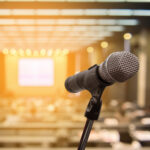 Close-up of a microphone on a stand in the foreground with a blurred auditorium and presentation screen in the background, suggesting a lecture or conference setting.