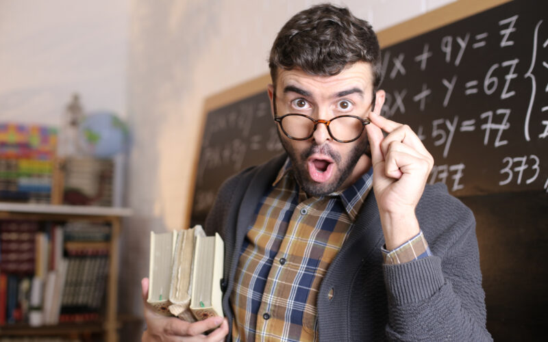 Alt text: A surprised-looking man wearing glasses holds two thick books in one arm and lifts his glasses with the other, standing in front of a chalkboard filled with math equations in a classroom.