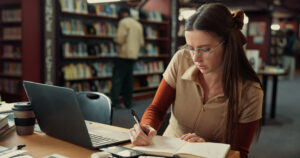 Student sits at table in library with laptop in front of her as she write in a notebook