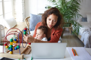 A mother sits at a laptop while holding a baby and guiding the child’s hand on a colorful bead maze toy at a home desk.