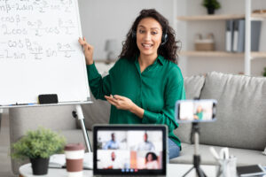 Professor wearing a headset presents math formulas on a whiteboard while conducting an online lesson with students visible on a laptop screen.