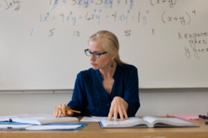 A professor reviews textbooks at a desk in front of a whiteboard covered with algebra equations.