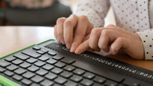 A person uses their fingertips to read text on an electronic braille display connected to a computer keyboard.