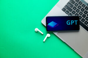 A smartphone displaying a GPT interface rests on a laptop beside wireless earbuds on a bright green surface.