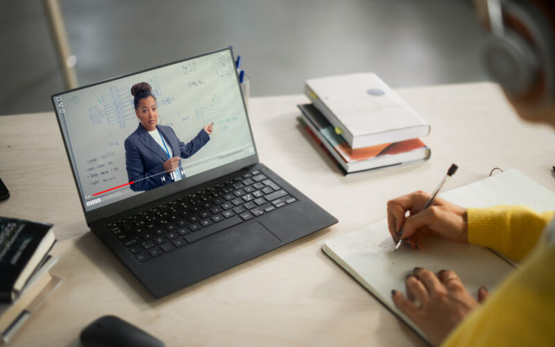A student wearing headphones takes notes in a notebook while watching an online lecture on a laptop where a teacher points to diagrams on a whiteboard.