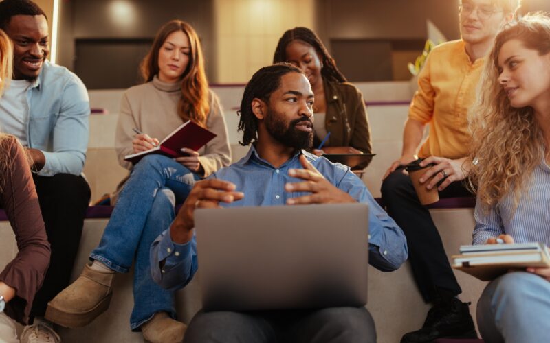 The image shows a college seminar setting. A professor with a beard sits in the foreground with a laptop, gesturing as he explains something to the group. Around him, college students sit on tiered seating, listening attentively, taking notes in notebooks, or holding tablets and coffee cups. The atmosphere is informal and collaborative, suggesting a discussion-based class rather than a lecture.