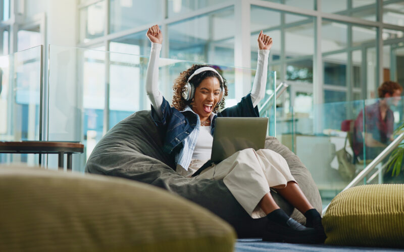 A woman wearing headphones sits on a beanbag chair with a laptop, raising her arms in excitement inside a modern, glass-walled building.