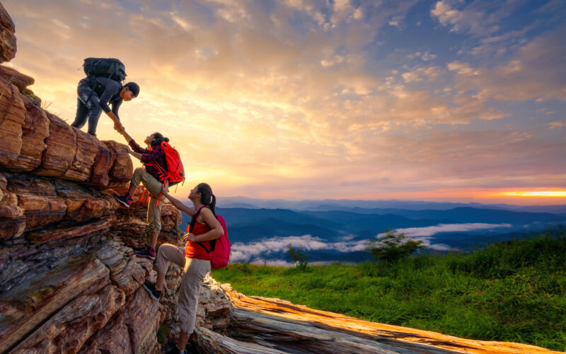 Three hikers climb a rocky outcrop at sunrise, with one person at the top pulling another up while a third steadies them from below; all wear backpacks, and a wide mountain landscape with layered hills and clouds stretches into the distance under a colorful sky.