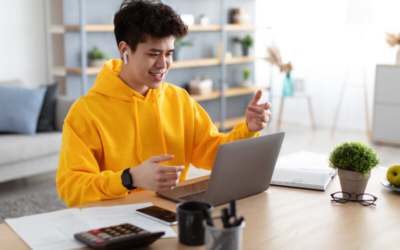 A person wearing a bright yellow hoodie sits at a desk using a laptop, smiling and gesturing with their hands as if recording a spoken response to a video. They are wearing wireless earbuds. On the desk are papers, a calculator, a smartphone, a notebook, a coffee mug, and a small potted plant. The background shows a softly lit living space with shelves and decorative items.