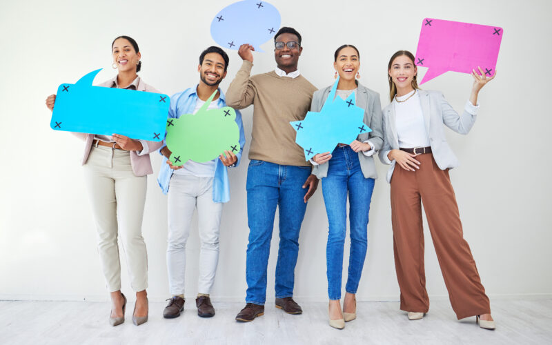 Five adults standing side by side against a plain background, smiling and holding colorful speech bubble signs in different shapes, suggesting conversation or discussion.