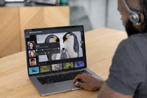 A man wearing headphones sits at a wooden table using a laptop that displays an online discussion interface with a voice comment, student profile images, and a slideshow of visual content.